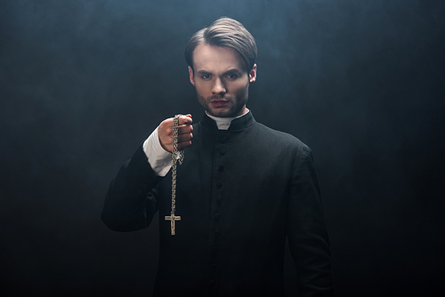 confident catholic priest holding necklace with cross while  on black background with smoke