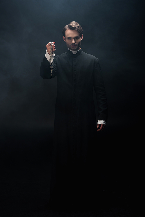 full length view of confident catholic priest holding necklace with cross on black background with smoke