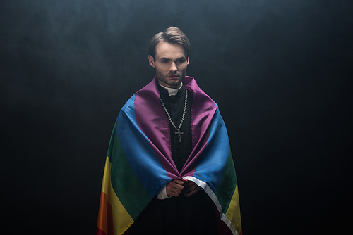serious catholic priest wrapped in lgbt flag while  on black background with smoke