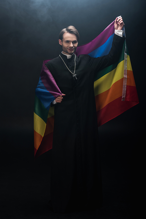 full length view of smiling catholic priest holding lgbt flag while  on black background with smoke