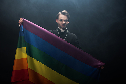 smiling catholic priest holding lgbt flag while  on black background with smoke