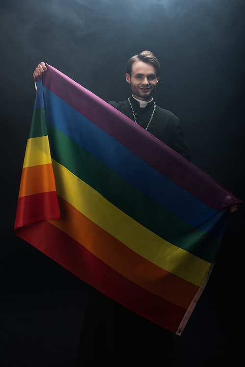 smiling catholic priest holding lgbt flag while  on black background with smoke