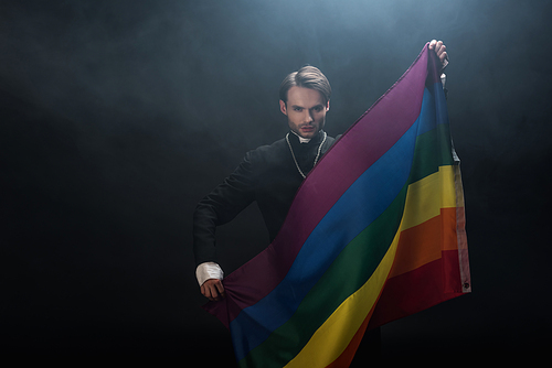 confident catholic priest holding lgbt flag while  on black background with smoke
