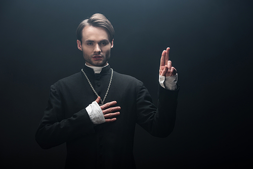 confident catholic priest showing blessing gesture and holding hand on chest isolated on black