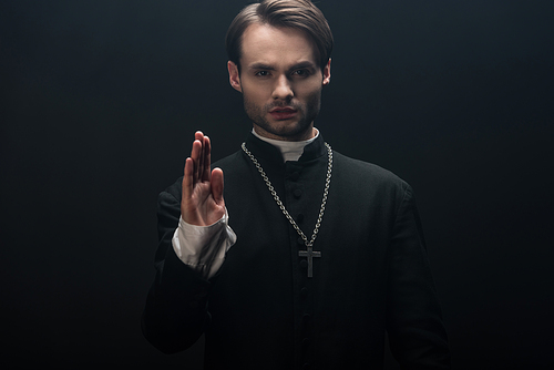 young confident catholic priest  and showing blessing gesture isolated on black