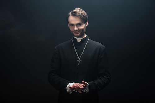 young smiling catholic priest standing with folded hands and  isolated on black