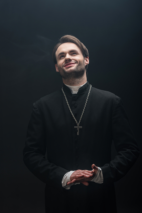 happy catholic priest standing with folded hands and looking up isolated on black