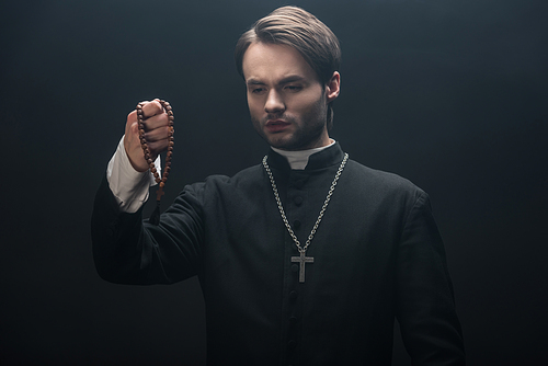 young tense catholic priest looking at wooden rosary beads isolated on black
