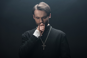 thoughtful catholic priest kissing silver cross with closed eyes isolated on black