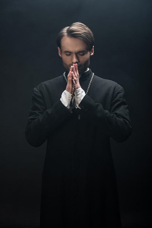 young concentrated catholic priest praying with closed eyes isolated on black