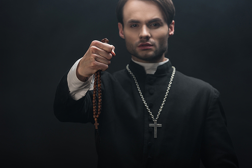 selective focus of young confident catholic priest holding wooded rosary beads in outstretched hand isolated on black