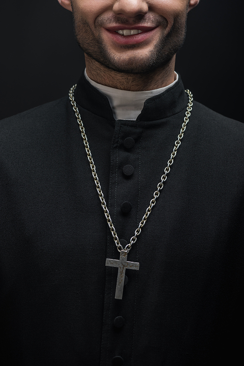 cropped view of smiling catholic priest with silver cross on necklace isolated on black