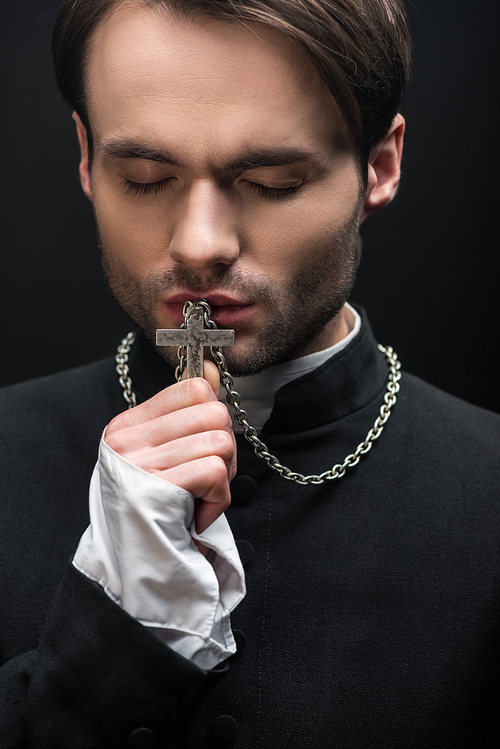 young thoughtful catholic priest kissing cross with closed eyes isolated on black
