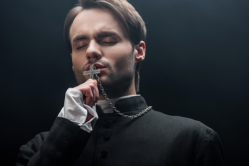 low angle view of young thoughtful catholic priest kissing cross with closed eyes isolated on black
