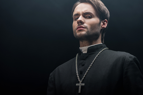low angle view of young tense catholic priest  isolated on black