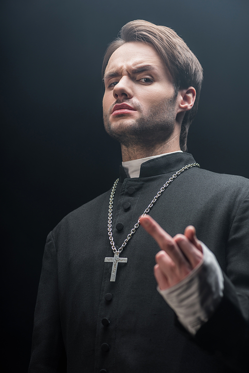 low angle view of dissatisfied catholic priest showing middle finger isolated on black