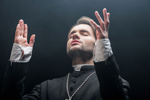 low angle view of young concentrated catholic priest praying with closed eyes isolated on black