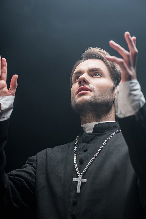 low angle view of young serious catholic priest praying with raised hands isolated on black