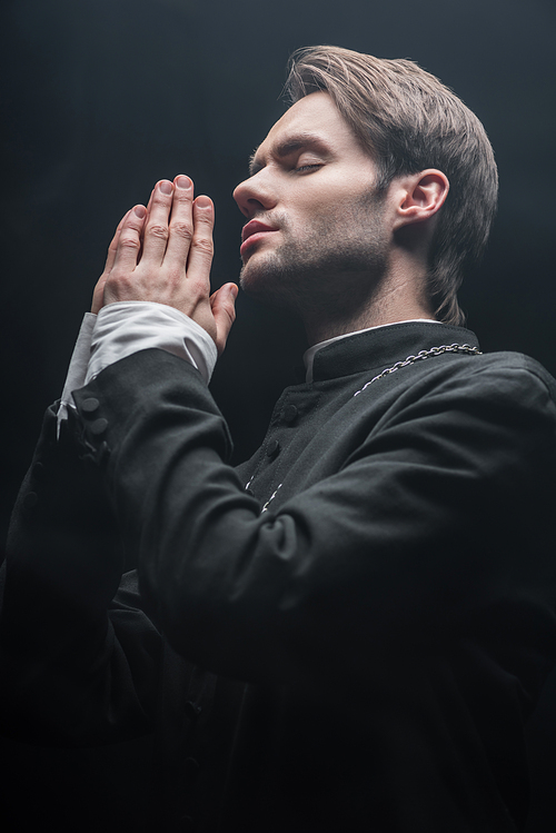 low angle veiw of young concentrated catholic priest praying with closed eyes isolated on black