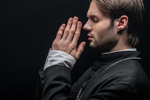 side view of young concentrated catholic priest praying with closed eyes isolated on black