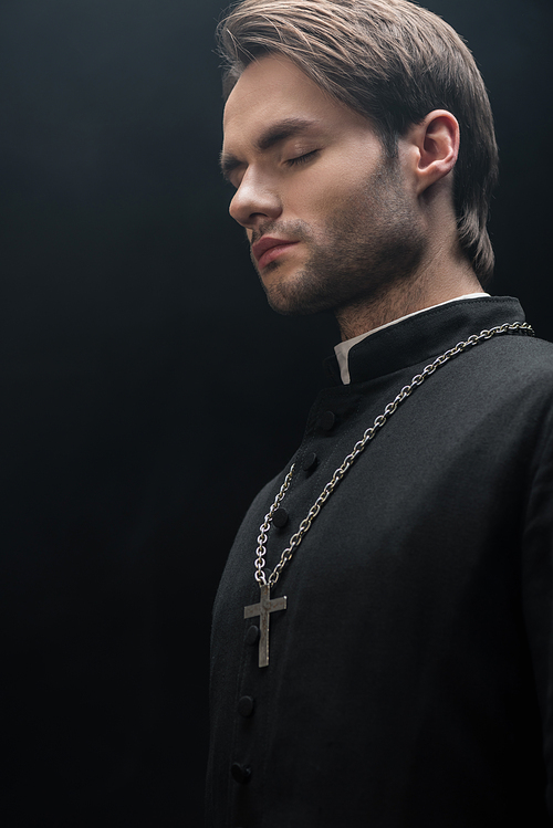 young catholic priest standing in darkness with closed eyes isolated on black