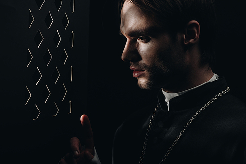 young tense catholic priest looking through confessional grille in dark with rays of light