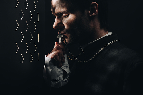 young tense catholic priest kissing cross on his necklace in dark near confessional grille with rays of light