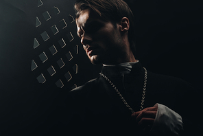 young serious catholic priest touching cross on his necklace in dark near confessional grille