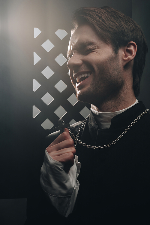sarcastic catholic priest laughing while touching cross on his necklace near confessional grille in dark with rays of light