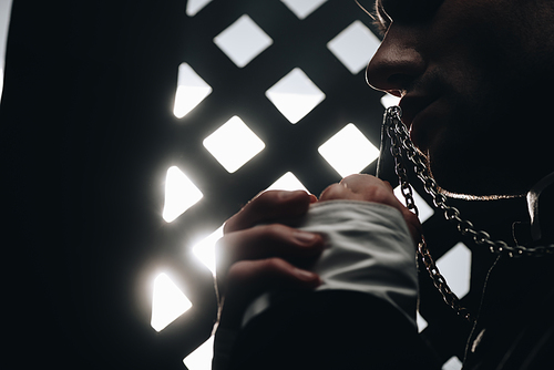 cropped view of catholic priest kissing cross on his necklace near confessional grille in dark with rays of light