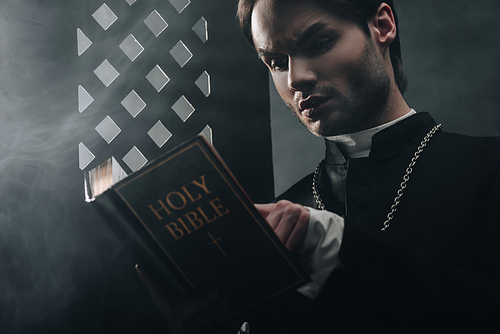 young thoughtful catholic priest reading bible near confessional grille in dark with rays of light