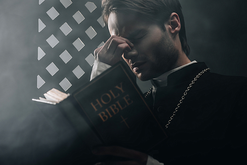 young thoughtful catholic priest touching face while reading bible near confessional grille in dark with rays of light