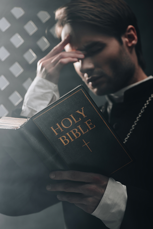 young serious catholic priest touching face while reading bible near confessional grille in dark with rays of light