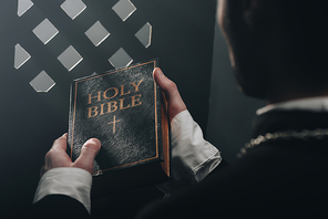 cropped view of catholic priest holding holy bible near confessional grille in dark with rays of light