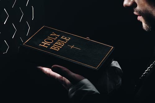 cropped view of catholic priest holding holy bible near confessional grille in dark with rays of light