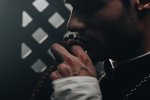 young catholic priest kissing wooden rosary beads near confessional grille in dark with rays of light