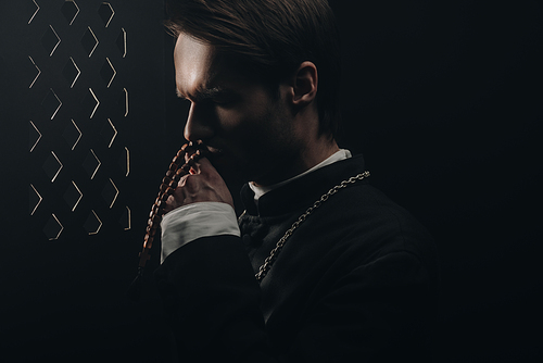 young thoughtful catholic priest kissing wooden rosary beads near confessional grille in dark with rays of light