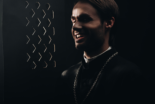 young sarcastic catholic priest laughing near confessional grille in dark with rays of light