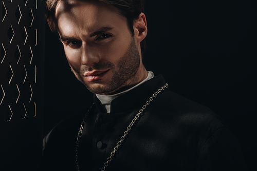 young confident catholic priest  near confessional grille in dark with rays of light