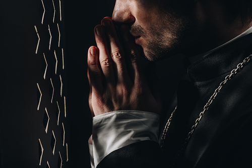 cropped view of catholic priest praying near confessional grille in dark with rays of light