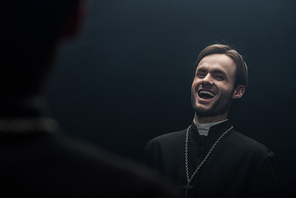 young catholic priest laughing sarcastically while looking at own reflection isolated on black