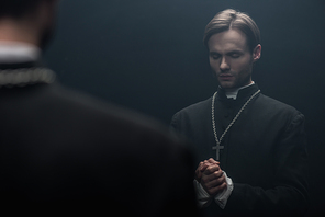 young serious catholic priest praying with closed eyes near own reflection isolated on black