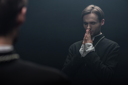 young serious catholic priest praying with closed eyes near own reflection isolated on black