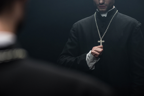 cropped view of catholic priest touching cross on his necklace near own reflection isolated on black