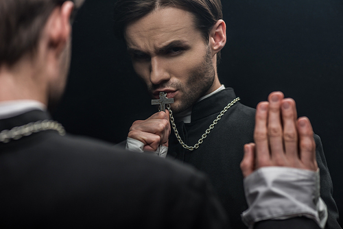 young tense catholic priest kissing holy cross while looking at own reflection isolated on black