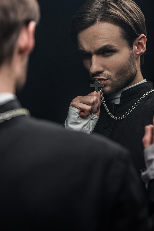 young tense catholic priest kissing holy cross while looking at own reflection isolated on black