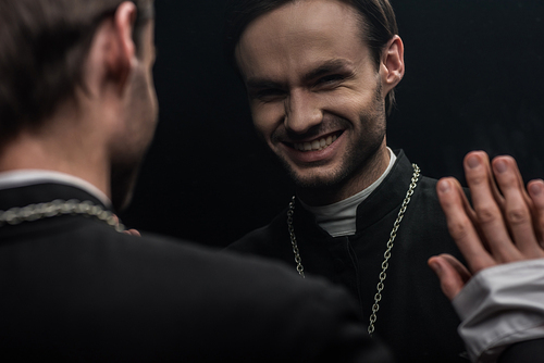 young catholic priest laughing sarcastically while looking at own reflection isolated on black