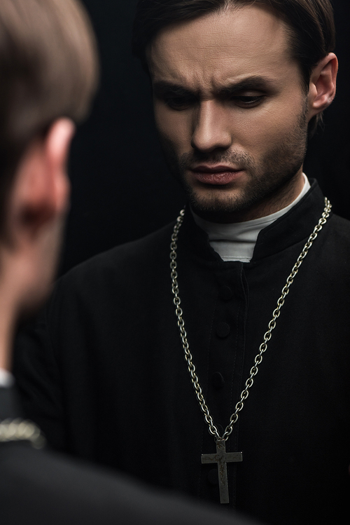 young serious catholic priest thinking while standing near own reflection isolated on black