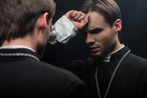young tense catholic priest holding fist near forehead while standing near own reflection isolated on black