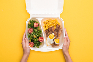 cropped view of woman holding  package with corn, meat, fried eggs and salad isolated on yellow